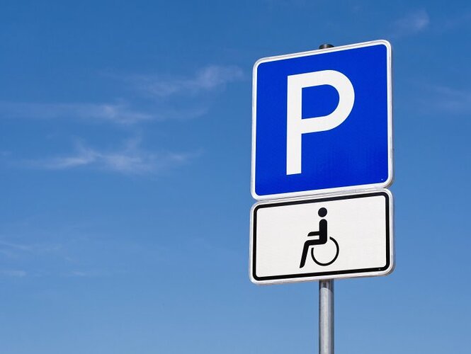 Ein blaues Verkehrsschild mit einem großen weißen P darauf. Darunter hängt ein weißes Verkehrsschild, auf welchem ein Rollstuhlfahrer abgebildet ist. Im Hintergrund ist blauer Himmel.