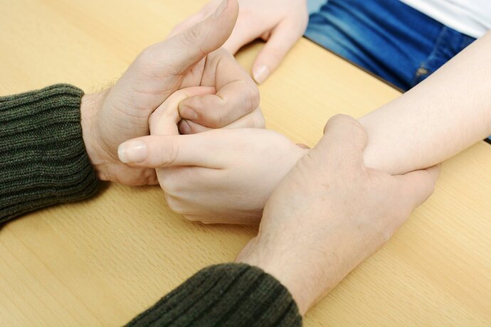 Eine Person hält mit der einen Hand das Handgelenk einer anderen Person fest. Mit der anderen Hand hat sie die Finger der Hand umschlossen. Die Hände liegen auf einem Tisch.