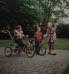 Auf dem Bild sind drei Kinder draußen zu sehen mit einem Skateboard, einem Fahrrad und einem Dreirrad mit Haltestange. Im Hintergrund sind Bäume.)