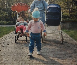 Im Vordergrund ist ein kleines Mädchen, welches auf Kopfsteinpflaster läuft. Im Hintergrund sind ein Kinderwagen und ein Rollstuhl mit einem Mädchen zu sehen.