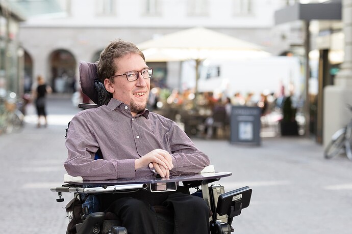 Ein Mann sitz in einem Elektro-Rollstuhl. Er lächelt in die Kamera. Im Hintergrund ist ein Markplatz zu sehen.