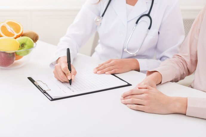 Eine Ärztin und eine Frau sitzen an einem Tisch. Die Ärztin hält einen Stift in der Hand und schreibt in ein Dokument auf einem Klemmbrett. Auf dem Tisch steht im Hintergrund eine Schale mit Obst.