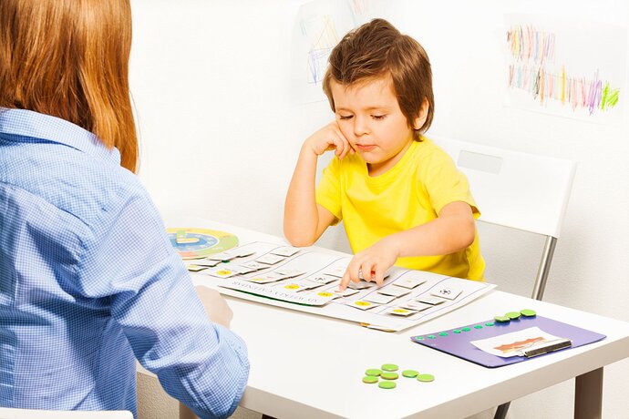 Ein kleiner Junge sitzt an einem Tisch. Vor ihm liegen verschiedene Karten und weitere Arbeitsmaterialien. Mit der einen Hand zeigt er auf ein Kärtchen und mit der anderen Hand stützt er seinen Kopf ab. Ihm gegenüber sitzt eine Frau