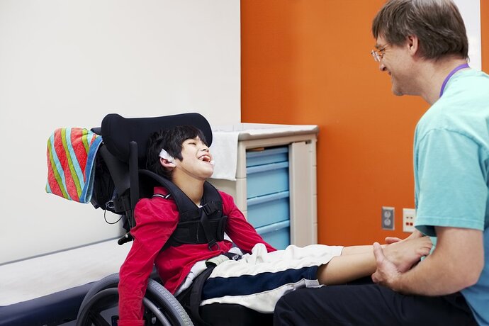 Ein Arzt sitzt einem Jungen im Rollstuhl in einem Behandlungszimmer gegenüber. Die Beine des Jungen liegen auf dem Schoß es Arztes. Beide lachen.