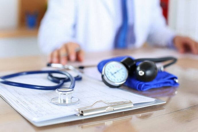 Auf einem Tisch liegen ein Stethoskop, ein Blutdruckmessgerät und eine Klemmbrett mit Papieren. Im Hintergrund ist ein Arzt zu sehen.
