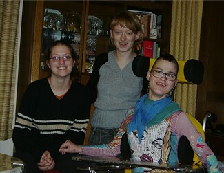 Drei junge Frauen in einem Wohnzimmer. Alle lächeln. Die Frau rechts auf dem Bild sitzt im Rollstuhl.