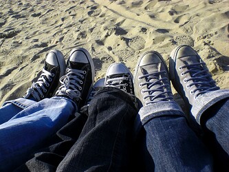 Im Vordergrund sind drei paar Schuhe mit Hosenbeinen. Im Hintergrund ist Sand.