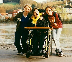 In der Mitte ist eine junge Frau im Rollstuhl. Links und rechts von ihr stehen zwei junge Frauen. Im Hintergrund ist ein Fluss.