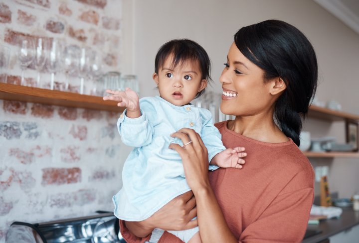 Eine glückliche Mutter hält ihr Baby mit Down-Syndrom auf dem Arm. Sie befinden sich in einer Küche in einem Einfamilienhaus.