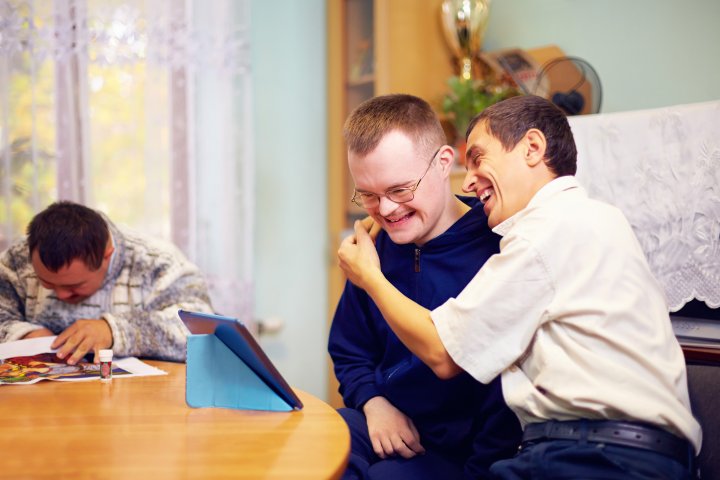 Zwei glückliche Freunde mit Behinderung, die sich umarmen, lachen und auf ein Tablet schauen.